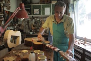 A guitar maker in an apron working on an acoustic guitar neck in a workshop, with the guitar body on the bench