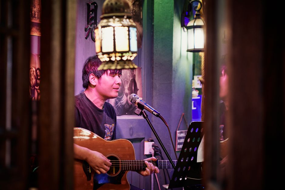 A musician playing acoustic guitar and singing into a microphone at a small indoor venue