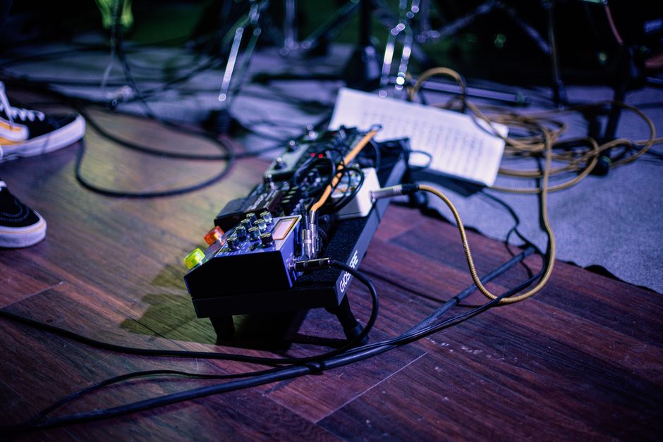 A pedalboard with cables and a setlist on a wooden stage floor