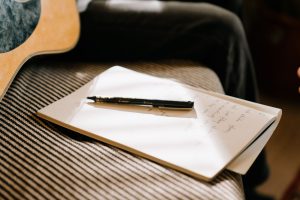 An open notepad with handwritten notes and a pen resting on it, with the edge of an acoustic guitar alongside